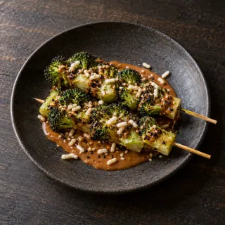 Two skewers of chargrilled broccoli stem and small florets on peanut mole, puffed rice and sesame