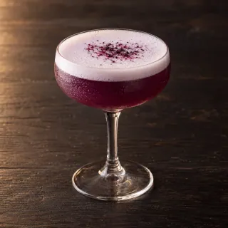 A deep violet purple-corn and pisco sour in a plain coupe glass with a thin white aquafaba foam top. A realistic small cocktail, no milk or egg props
