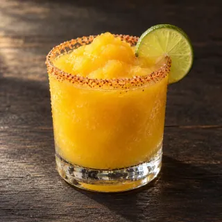 A vivid golden frozen mango, rum and lime sour with a naturally slushy texture in a simple short clear glass, a small lime wheel and a light chilli-salt rim