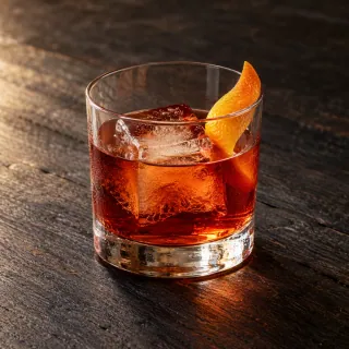 An amber-red gin, plum, bitter orange and vermouth Negroni in an unbranded clear short rocks glass with one large ice cube and a twist of orange peel