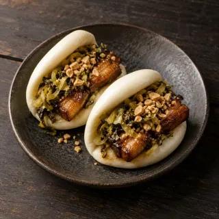 Two fluffy folded steamed bao buns containing glazed pork belly, pickled mustard greens and crushed peanut