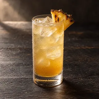 An amber golden roasted pineapple, ginger and soda drink in a clear highball glass with realistic ice and a small charred pineapple wedge. No branded bottle or alcohol