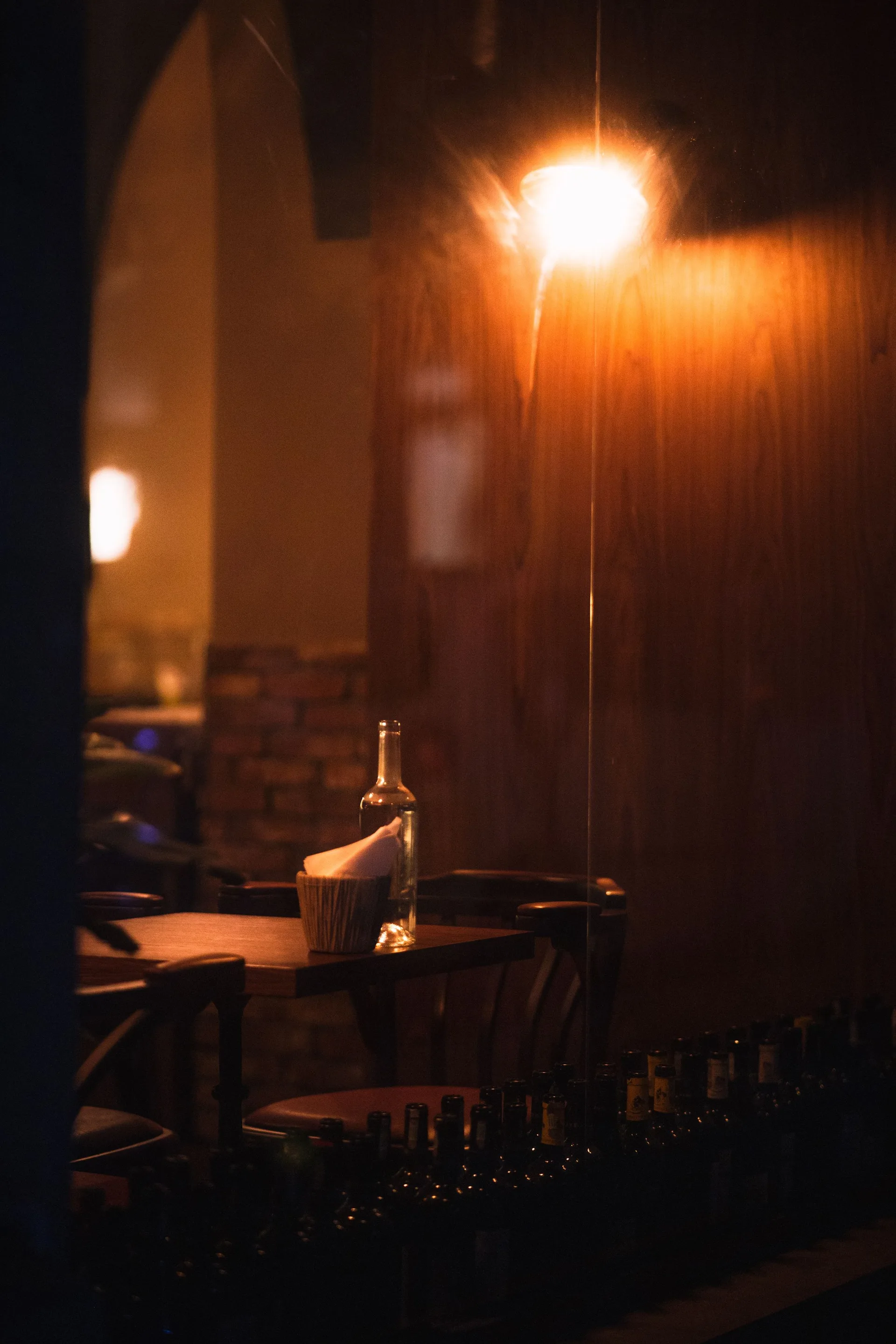 A dimly lit bar table with a single bottle, warm light spilling across dark wood