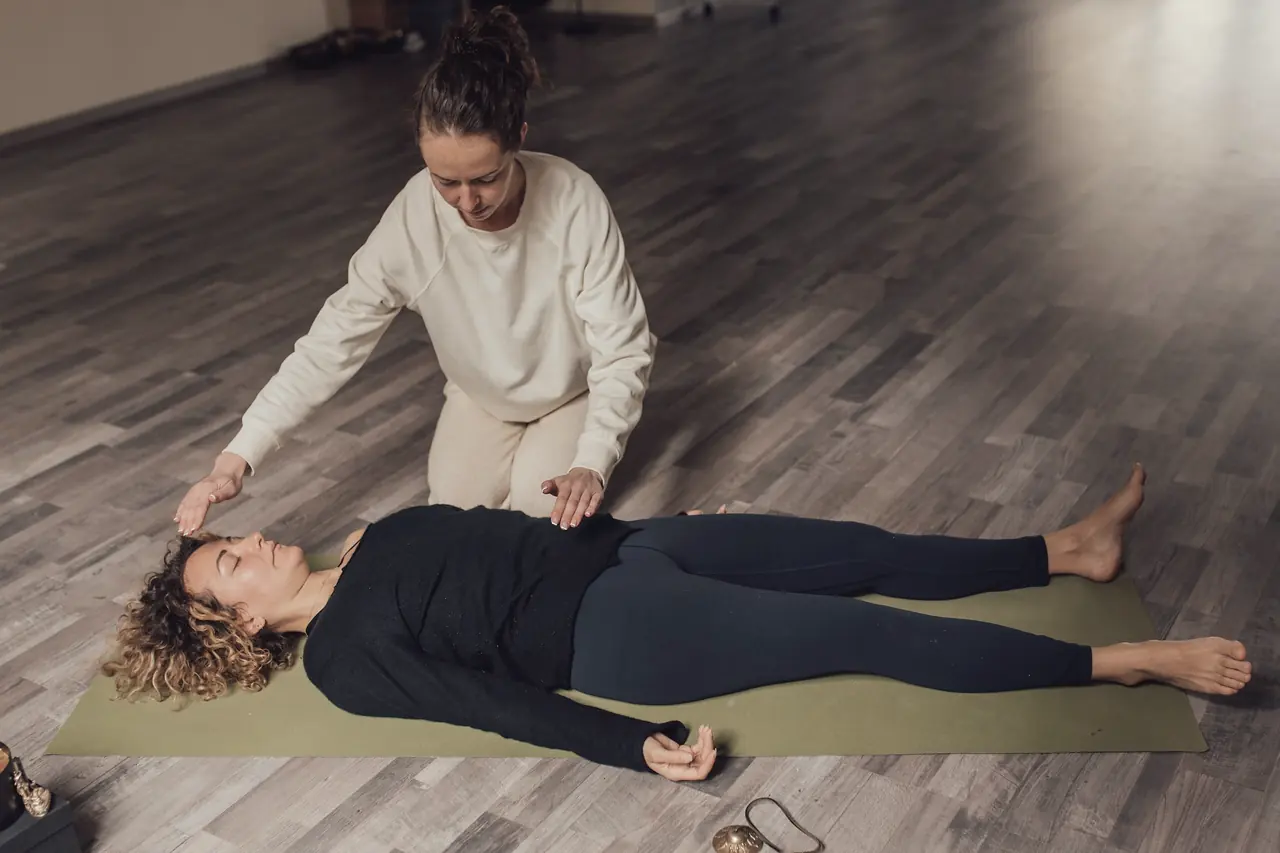 A student lying in savasana on a warm wooden floor while a teacher stands quietly at her head