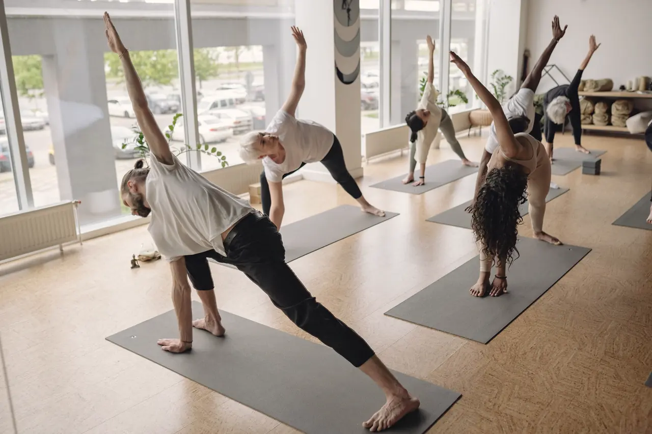 A class holding triangle pose in a row beside tall windows