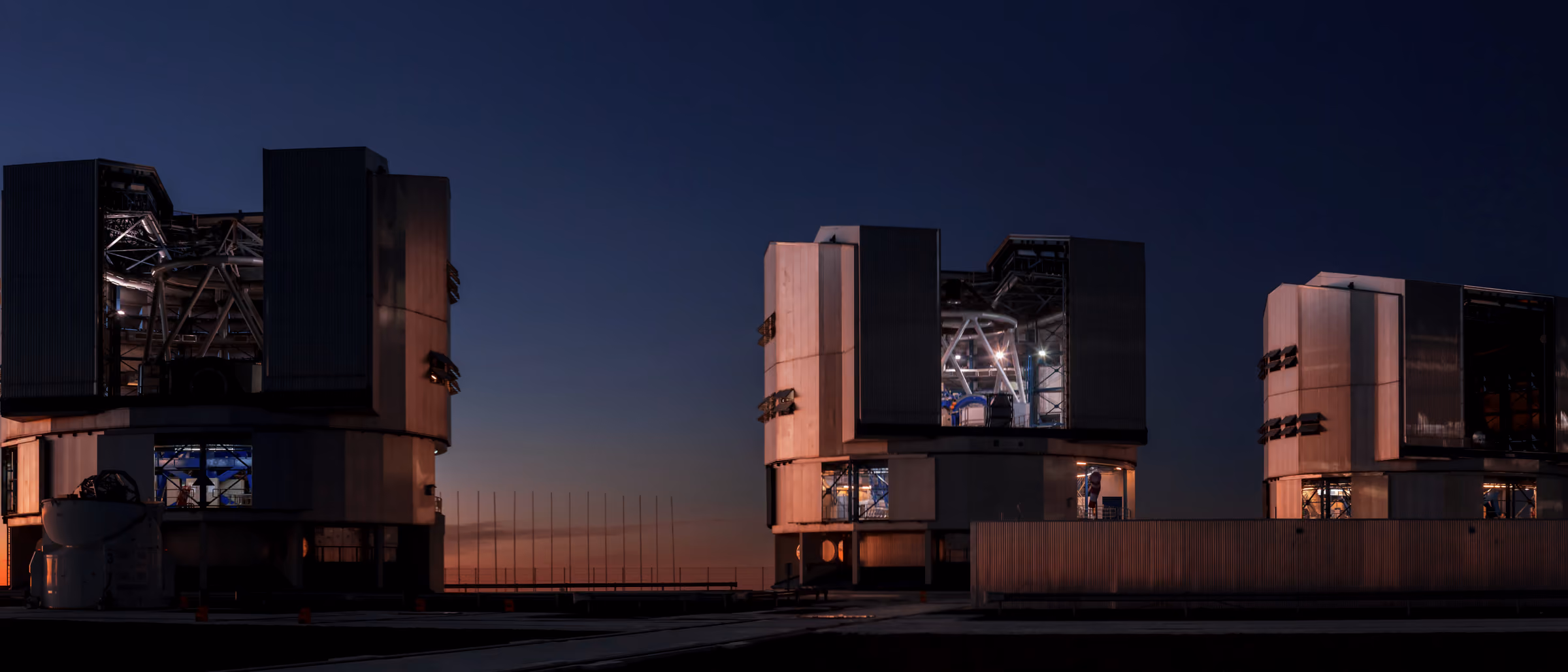 Observatory domes on a flat desert platform against a deep ochre sunset, two figures silhouetted at the edge.