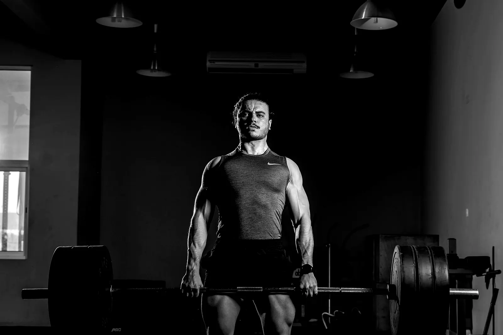 A lifter standing with a loaded barbell held at the hips in a dark studio, the room falling away behind them
